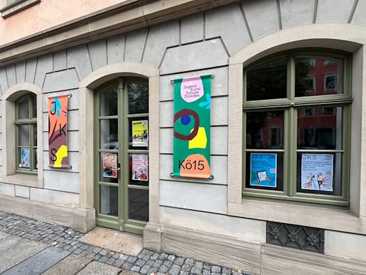 Erdgeschossfassade eines Altbaus in Dresden mit grün gestrichenen Fenstern und Türen der JugendKunstSchule. An der Wand hängen bunte Banner der Einrichtung „Kö15“, in den Scheiben kleben Informationsplakate für Kurse.