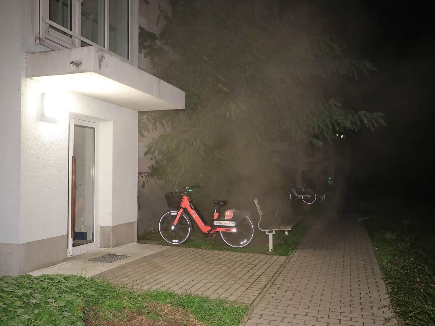 Nächtlicher Blick auf den Eingangsbereich eines Wohnblocks mit heller Beleuchtung; im dichten, wabernden Rauch an der Fassade sind ein rot-schwarzes Leihfahrrad und eine Parkbank zu erkennen.