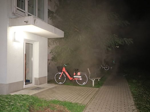 Nächtlicher Blick auf den Eingangsbereich eines Wohnblocks mit heller Beleuchtung; im dichten, wabernden Rauch an der Fassade sind ein rot-schwarzes Leihfahrrad und eine Parkbank zu erkennen.