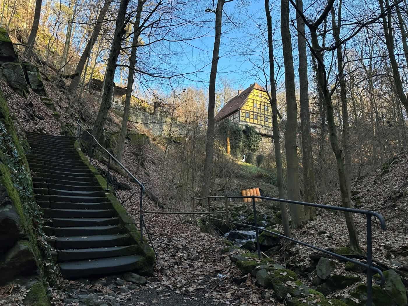 Blick in den waldigen Keppgrund mit einer alten, bemoosten Steintreppe samt Handlauf links und einem kleinen Bachlauf rechts; im Hintergrund thront ein historisches Fachwerkhaus auf dem Hang.