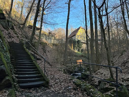 Blick in den waldigen Keppgrund mit einer alten, bemoosten Steintreppe samt Handlauf links und einem kleinen Bachlauf rechts; im Hintergrund thront ein historisches Fachwerkhaus auf dem Hang.
