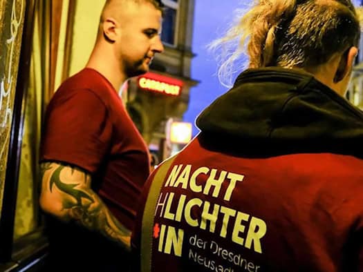 Zwei Mitarbeiter der Kiezlichter im nächtlichen Außeneinsatz; im Vordergrund ist die Rückansicht einer Person in dunkelroter Kleidung mit der gelben Aufschrift „Nachtlichter“ zu sehen.