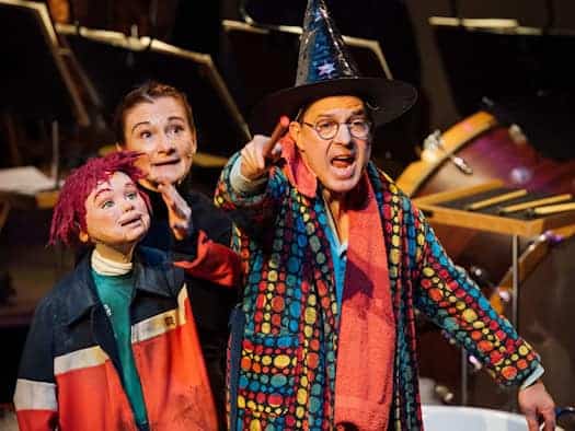 Szene des Kinderkonzerts „Carl und Caspar“ in der Semperoper: Ein Mann im bunt gepunkteten Bademantel mit Zauberhut zeigt mit einem Zauberstab nach vorne. Neben ihm steht eine Frau mit einer rothaarigen Puppe vor den Instrumenten des Orchesters.
