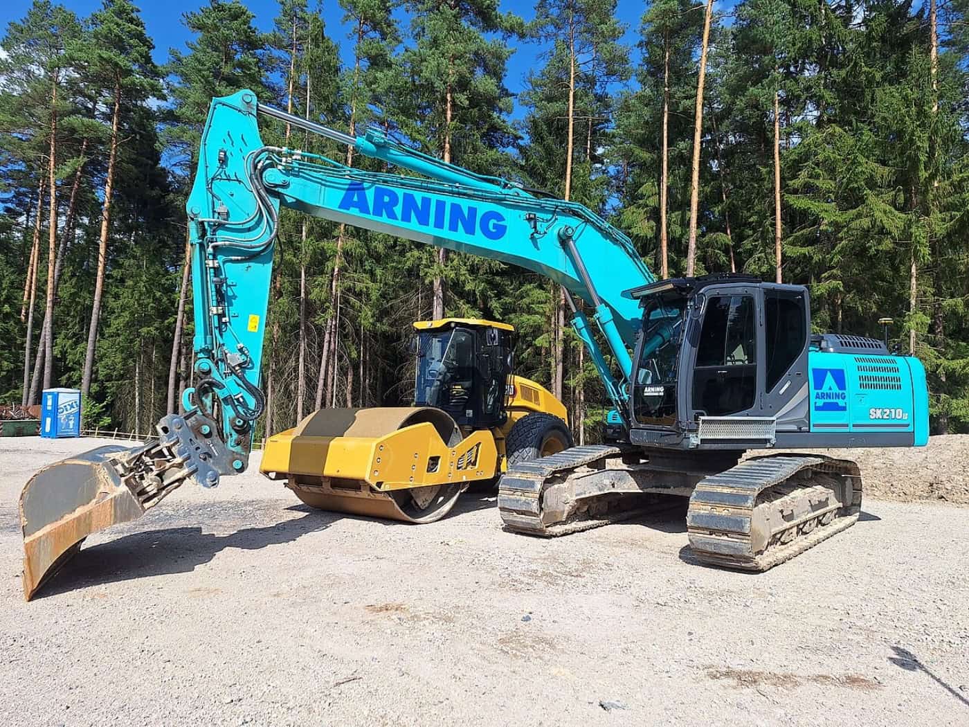 Türkisfarbener Kettenbagger und gelbe Straßenwalze auf einer Baustelle vor einem Wald.