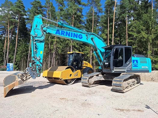 Türkisfarbener Kettenbagger und gelbe Straßenwalze auf einer Baustelle vor einem Wald.