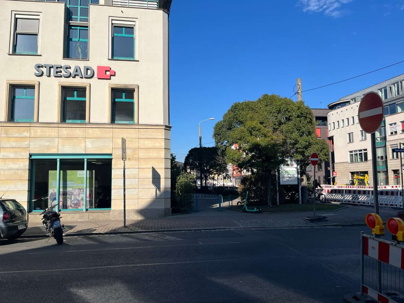Straßenansicht der Königsbrücker Straße in Dresden mit dem Gebäude der STESAD GmbH auf der linken Seite, davor parkende Fahrzeuge; rechts im Hintergrund sind Baustellenabsperrungen und Schilder unter blauem Himmel zu sehen.
