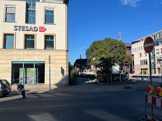 Straßenansicht der Königsbrücker Straße in Dresden mit dem Gebäude der STESAD GmbH auf der linken Seite, davor parkende Fahrzeuge; rechts im Hintergrund sind Baustellenabsperrungen und Schilder unter blauem Himmel zu sehen.