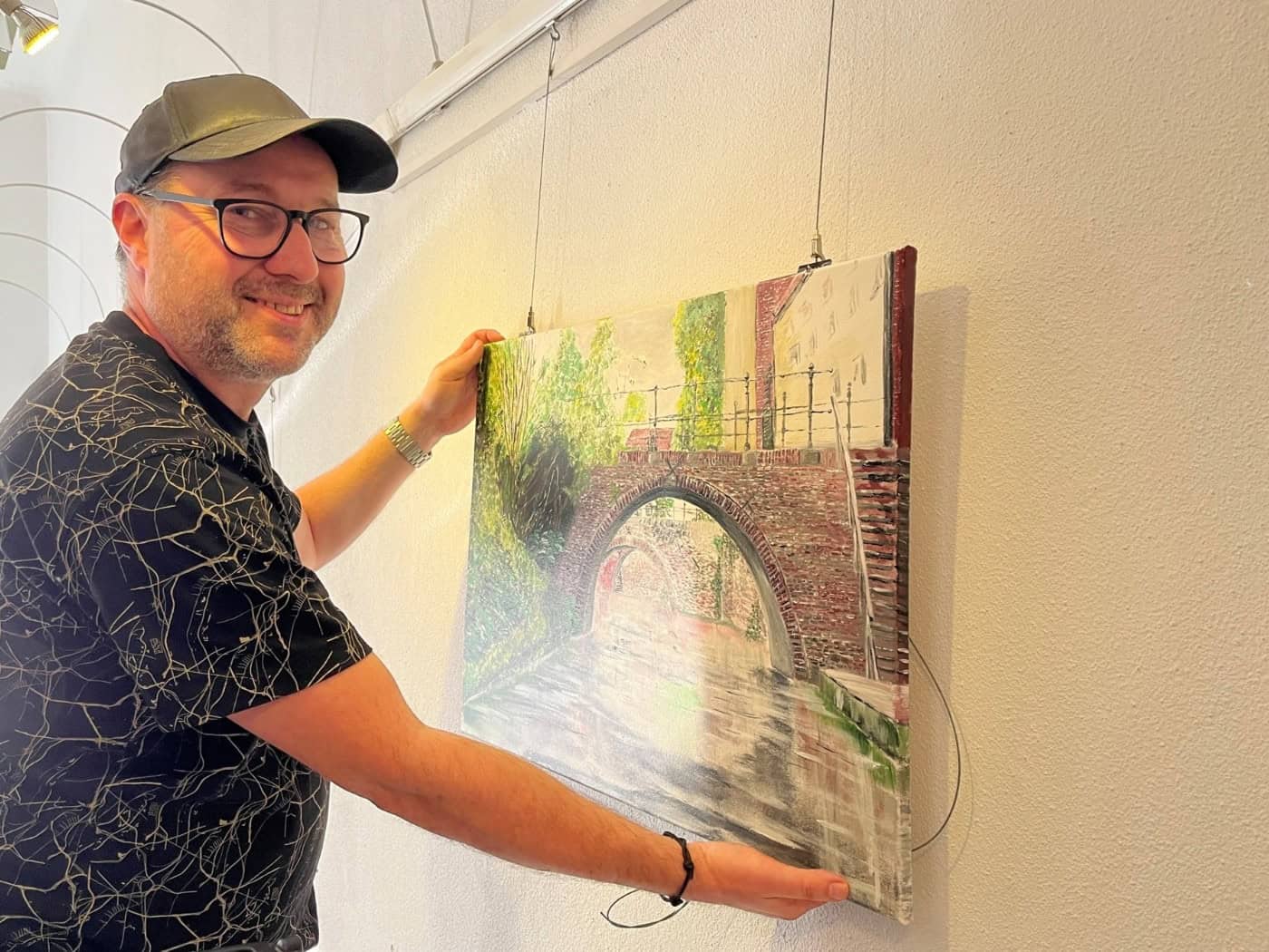 Mann mit Basecap und Brille hängt ein Acrylgemälde mit einer Backsteinbrücke an eine weiße Wand
