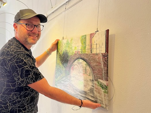 Mann mit Basecap und Brille hängt ein Acrylgemälde mit einer Backsteinbrücke an eine weiße Wand