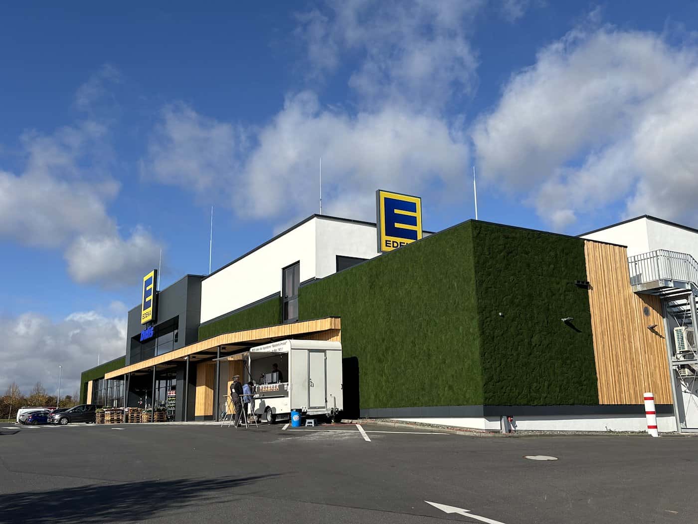 Außenansicht eines modernen EDEKA-Einkaufsmarktes unter blauem Himmel. Eine große Außenwand des Gebäudes ist komplett flächig mit sattgrünem Moos verkleidet und mit vertikalen Hölzern sowie EDEKA-Logos kombiniert.