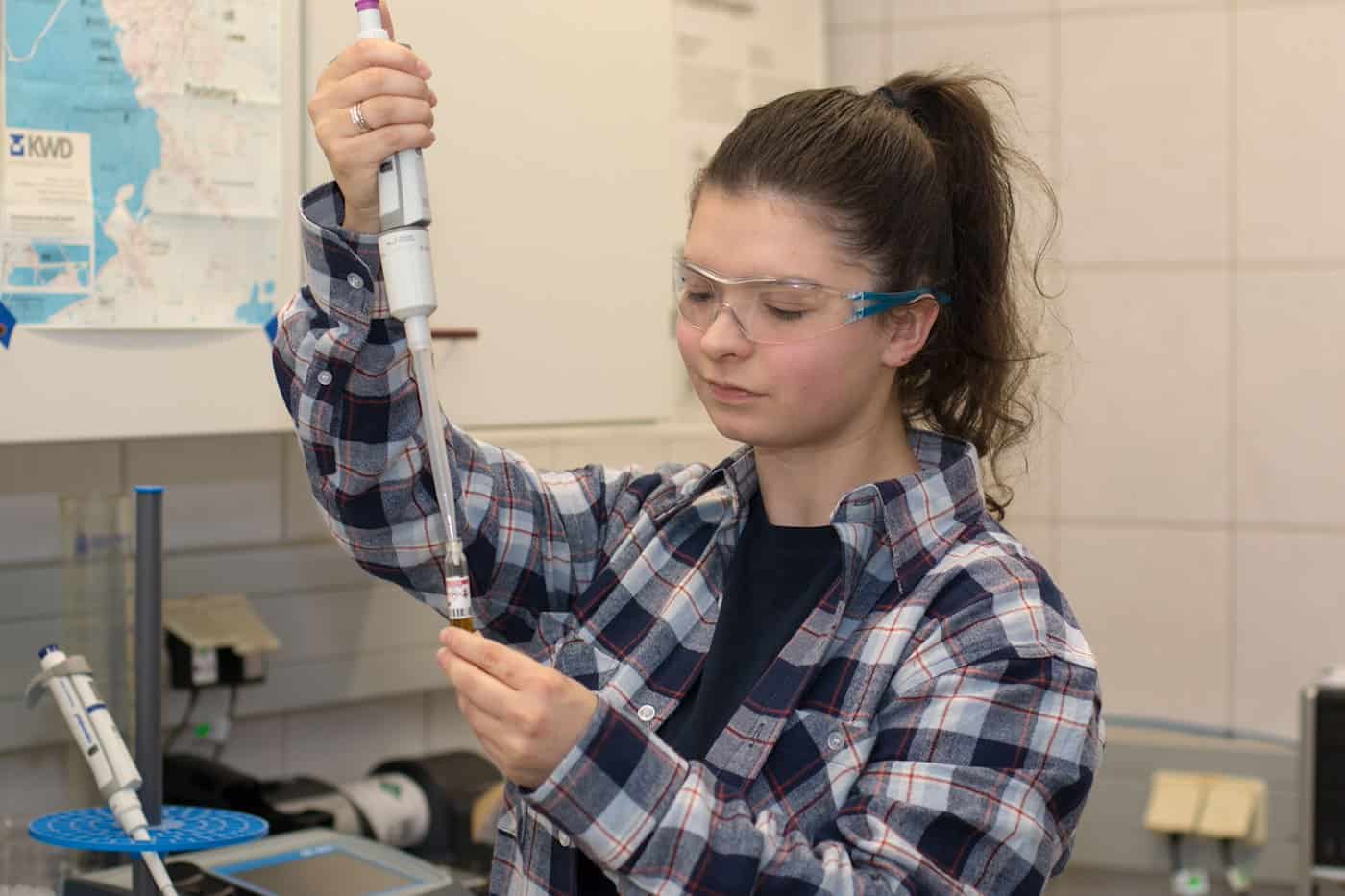Eine junge Frau mit Schutzbrille und kariertem Hemd zieht mit einer Dosierpipette eine Flüssigkeit aus einer kleinen Glasampulle im Labor. Im Hintergrund hängt eine Übersichtskarte an der gefliesten Wand.