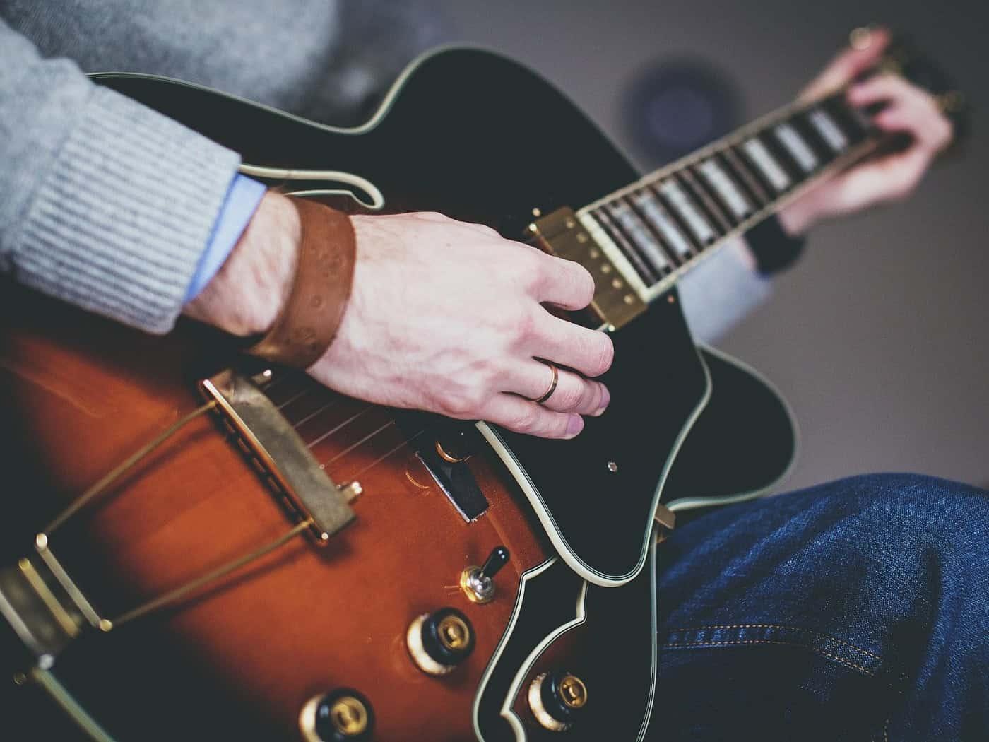 Symbolbild: Nahaufnahme der Hände eines Mannes in grauem Pullover, der die Saiten einer halbakustischen Gitarre greift.