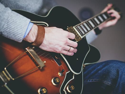 Symbolbild: Nahaufnahme der Hände eines Mannes in grauem Pullover, der die Saiten einer halbakustischen Gitarre greift.