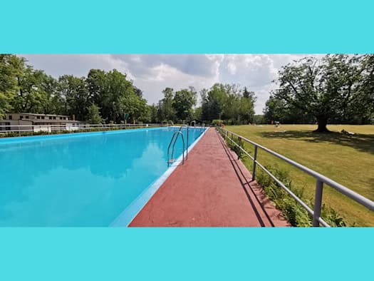 Blick über das leuchtend blaue, ungenutzte Schwimmbecken und den rot getuschten Beckenrand im Luftbad Dölzschen, flankiert von einer weitläufigen Liegewiese mit Bäumen unter wolkigem Himmel.