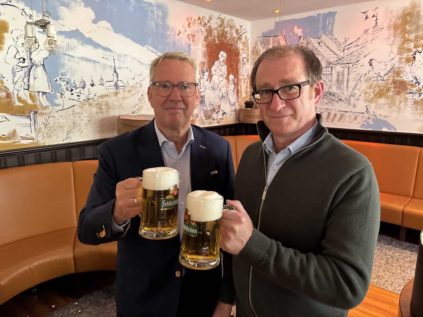 Uwe Tschuschke und Matthias Singer lächeln in die Kamera und stoßen im Restaurant mit zwei großen Krügen Feldschlößchen-Bier an.