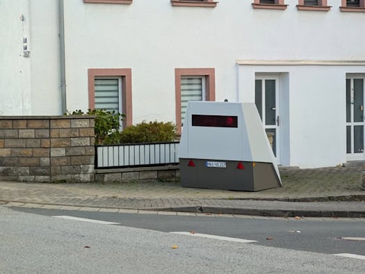 Nahaufnahme des grauen mobilen Blitzer-Anhängers mit der Kennung „HWI-VE 217“, dessen roter Infrarotblitz in der dunklen Sensoreinheit zu erkennen ist; im Hintergrund eine weiße Hauswand mit Fenstern.