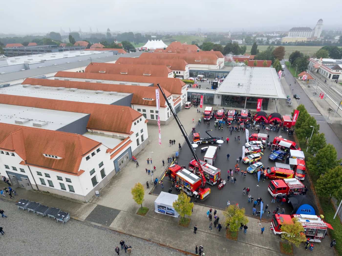 Luftaufnahme des Messegeländes Dresden: Auf dem Außengelände zwischen den Hallen stehen zahlreiche rote Feuerwehrfahrzeuge, Einsatzwagen und ein hoher Kran. Besucher gehen über das Gelände der Fachmesse FLORIAN.