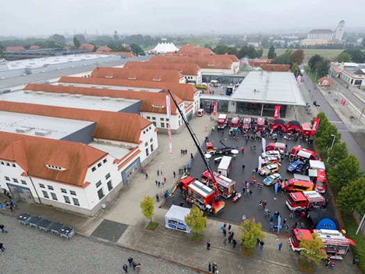 Luftaufnahme des Messegeländes Dresden: Auf dem Außengelände zwischen den Hallen stehen zahlreiche rote Feuerwehrfahrzeuge, Einsatzwagen und ein hoher Kran. Besucher gehen über das Gelände der Fachmesse FLORIAN.