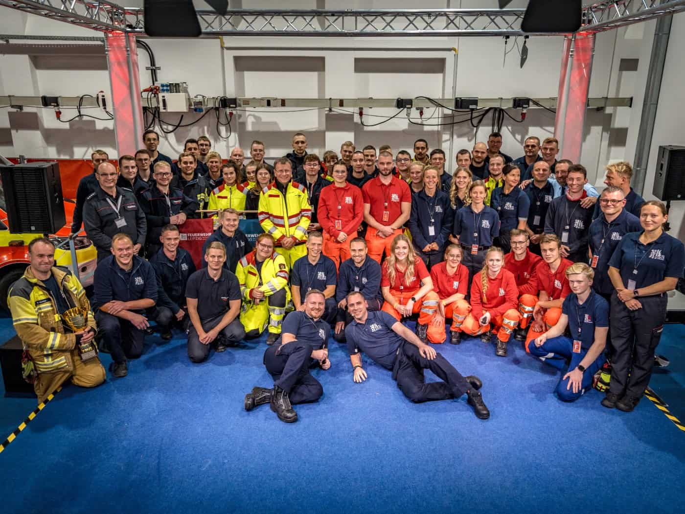 Gruppenfoto zahlreicher Einsatzkräfte von Feuerwehr und Rettungsdienst in Arbeitsbekleidung in Messehalle 4. Die Teams stehen, knien und liegen lächelnd auf blauem Messeboden vor einer Traverse und einem Einsatzfahrzeug.