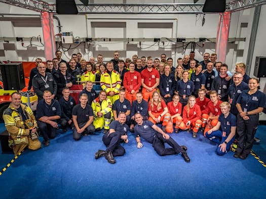 Gruppenfoto zahlreicher Einsatzkräfte von Feuerwehr und Rettungsdienst in Arbeitsbekleidung in Messehalle 4. Die Teams stehen, knien und liegen lächelnd auf blauem Messeboden vor einer Traverse und einem Einsatzfahrzeug.