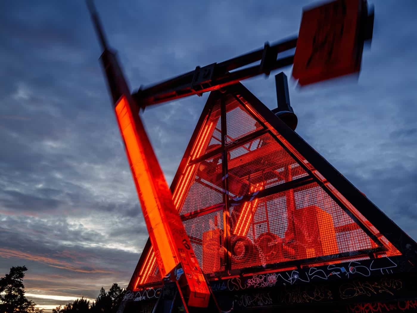 Rot beleuchtetes Prager Metronom mit schwingendem Pendel vor dem bewölkten Abendhimmel.