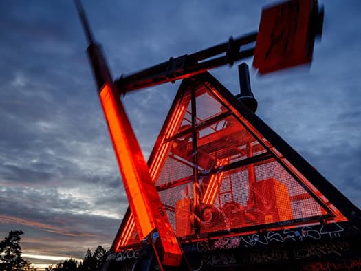 Rot beleuchtetes Prager Metronom mit schwingendem Pendel vor dem bewölkten Abendhimmel.