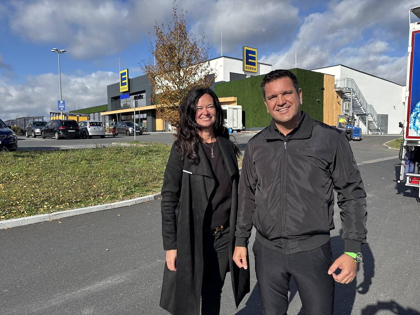 Lydia Förster und Andreas Bergermann stehen lächelnd nebeneinander auf dem Parkplatz vor dem EDEKA-Markt in Kürnach. Im Hintergrund ist die große grüne Moosfassade des Supermarktes gut zu erkennen.   SEO-gerechter Dateiname: