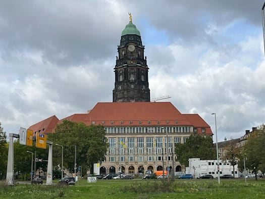 Außenansicht des Neuen Rathauses in Dresden mit seinem prägnanten Rathausturm und dem goldenen Rathausmann unter bewölktem Himmel, davor befinden sich Wiesenflächen und Verkehrsschilder.