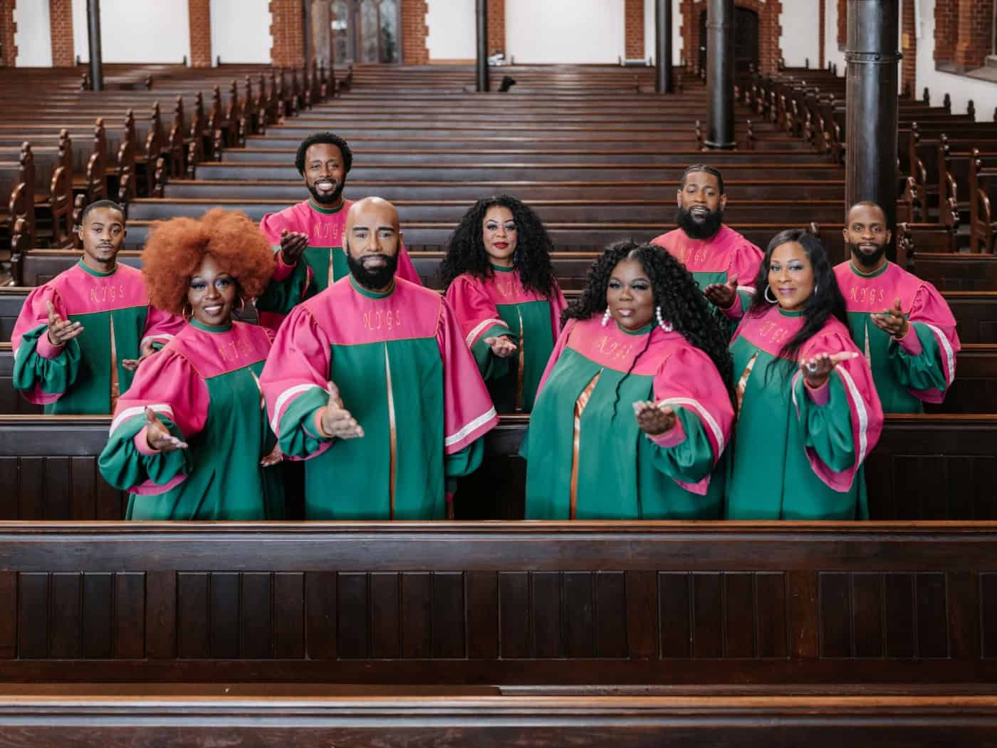 Acht Mitglieder des US-Chors New York Gospel Stars stehen lächelnd in leuchtend grün-pinken Roben zwischen den Holzbänken einer Kirche und strecken einladend die Hände nach vorne aus.