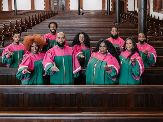 Acht Mitglieder des US-Chors New York Gospel Stars stehen lächelnd in leuchtend grün-pinken Roben zwischen den Holzbänken einer Kirche und strecken einladend die Hände nach vorne aus.