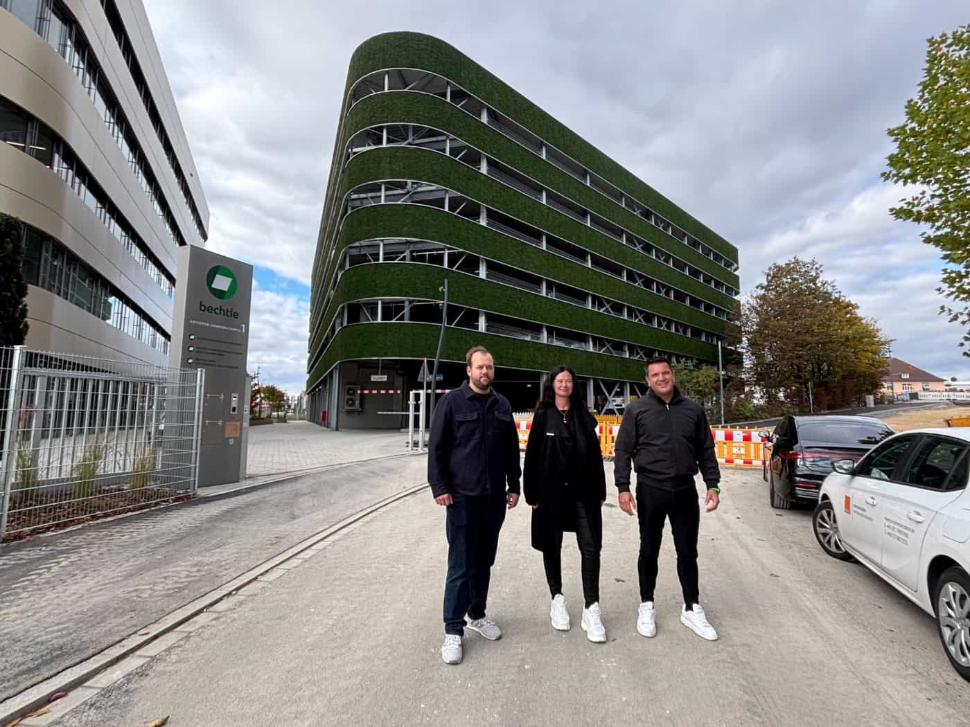Raphael Marquardt, Lydia Förster und Andreas Bergermann stehen vor einem mehrstöckigen, rund geschwungenen Parkhaus mit grüner Moosverkleidung an den Außenrängen. Links steht ein Firmenstele mit der Aufschrift „bechtle“.