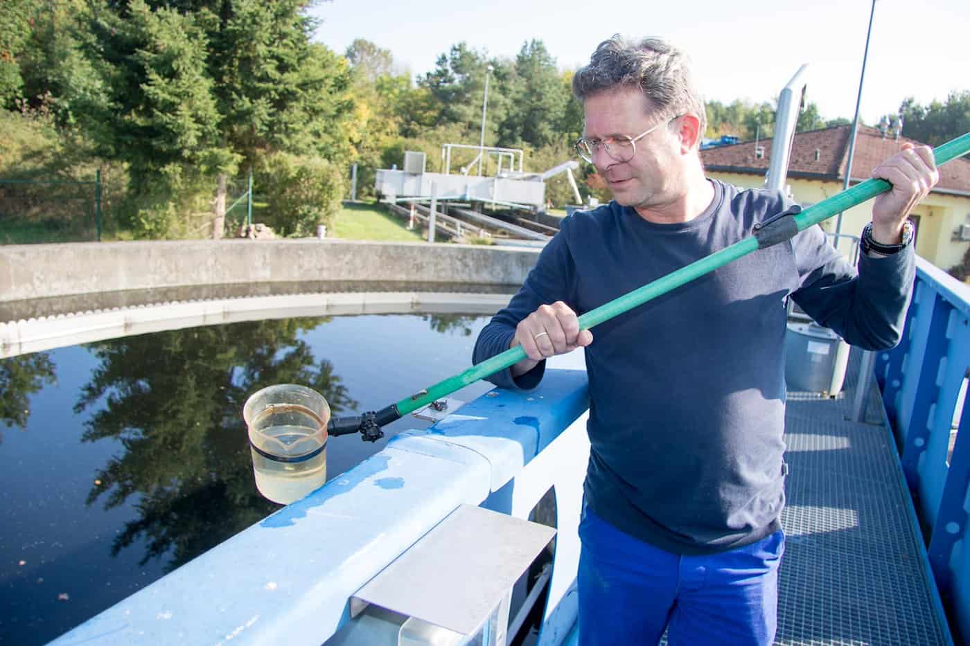 Ein Mann mit Brille lehnt an einem blauen Geländer und hält eine lange Teleskopstange mit einem transparenten Messbecher über ein rundes Nachklärbecken, um eine Wasserprobe zu nehmen.