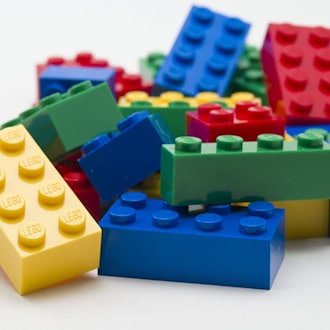Lose LEGO-Steine in Rot, Gelb, Grün und Blau auf einer hellen Fläche.