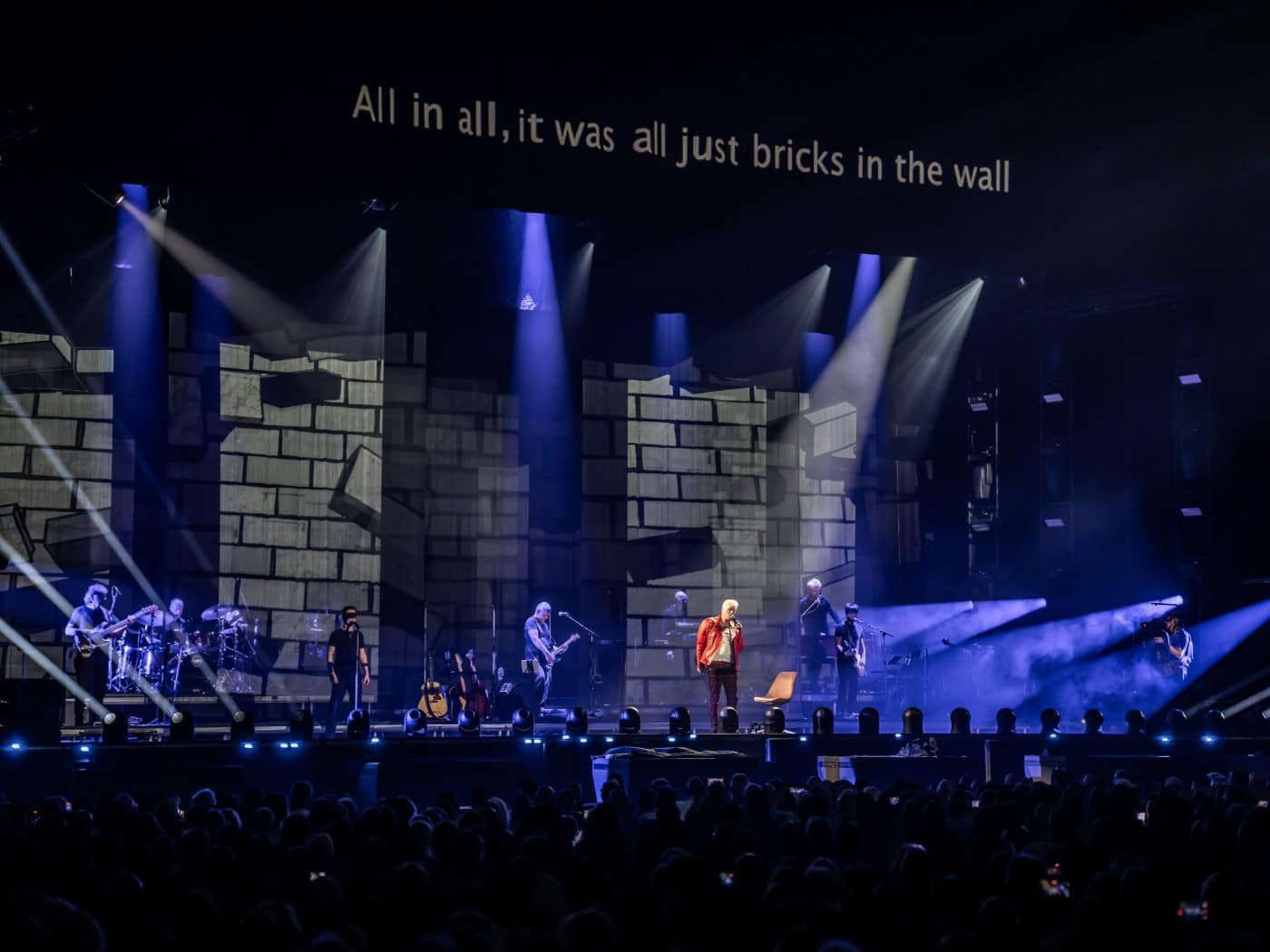 Bühnenansicht eines Konzerts des Pink Floyd Projects mit einer Kulisse aus gemalten Mauersteinen, Scheinwerferkegeln in blauem Licht und der Textzeile „All in all, it was all just bricks in the wall“ auf einer Leinwand oberhalb der Band.