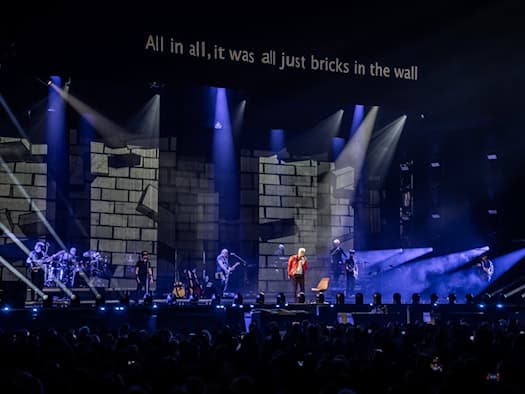 Bühnenansicht eines Konzerts des Pink Floyd Projects mit einer Kulisse aus gemalten Mauersteinen, Scheinwerferkegeln in blauem Licht und der Textzeile „All in all, it was all just bricks in the wall“ auf einer Leinwand oberhalb der Band.