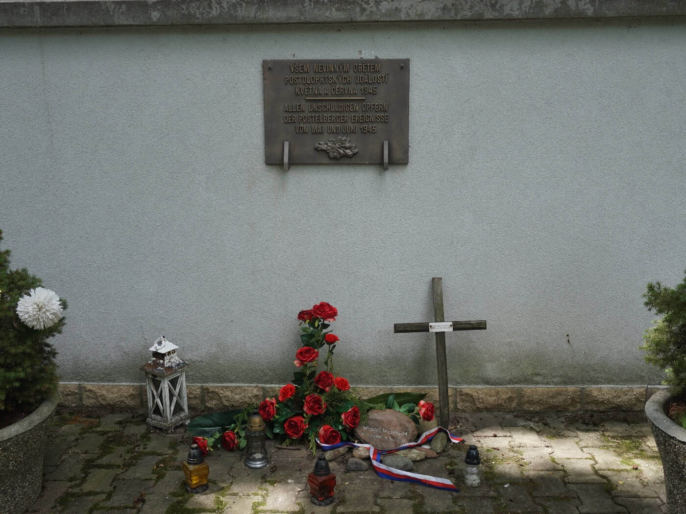 Deutsch-tschechische Gedenktafel in Postoloprty mit Blumen, Kerzen und einem Holzkreuz.