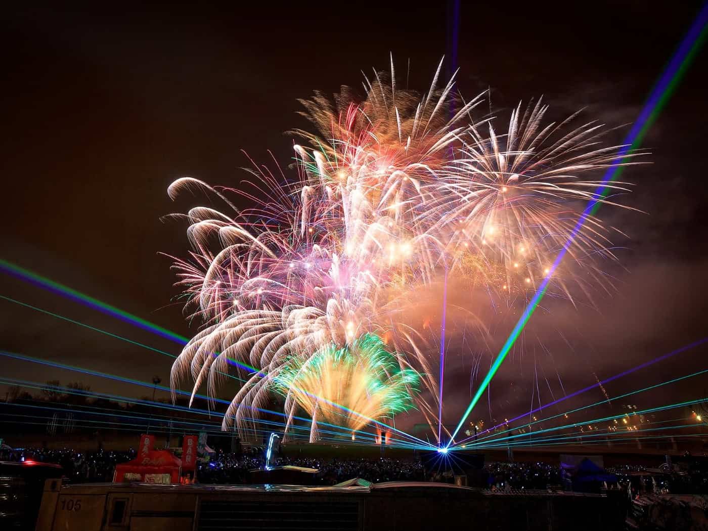 Farbenfrohes Feuerwerk und leuchtend grüne sowie blaue Laserstrahlen erhellen den nächtlichen Himmel über einer Freiluft-Veranstaltung. Im dunklen Vordergrund ist die Silhouette des Geländes mit Eventtechnik und Zuschauern zu erkennen.