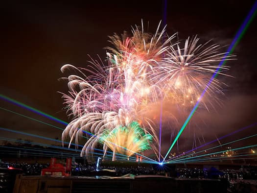 Farbenfrohes Feuerwerk und leuchtend grüne sowie blaue Laserstrahlen erhellen den nächtlichen Himmel über einer Freiluft-Veranstaltung. Im dunklen Vordergrund ist die Silhouette des Geländes mit Eventtechnik und Zuschauern zu erkennen.
