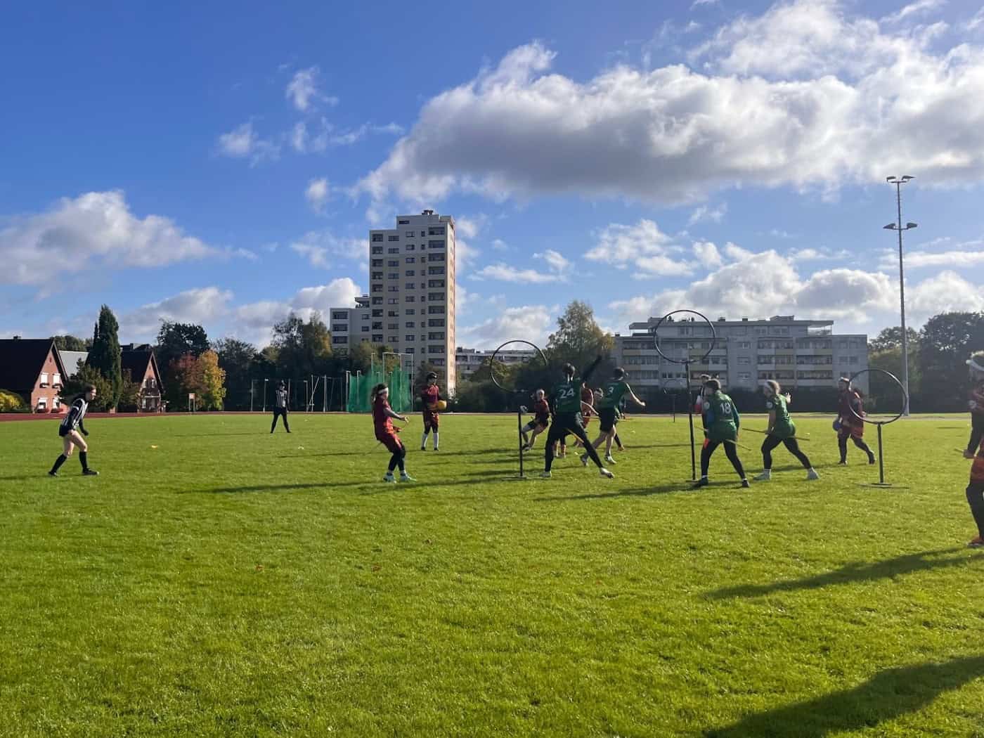 Quadball-Spieler auf einem grünen Sportplatz bei Sonnenschein; Teams laufen mit Plastikstangen zwischen den Beinen auf Tore aus Ring-Ständern zu. Im Hintergrund stehen Wohngebäude und Bäume unter blauem Himmel mit Schönwetterwolken.