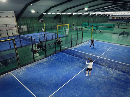 Blick von oben in eine große Sporthalle mit zwei blauen Indoor-Padel-Courts hinter Zäunen und Glaswänden. Personen spielen aktiv Padel-Tennis auf dem blauen Kunstrasen, im Hintergrund sind weitere Sportfelder der Halle zu sehen.