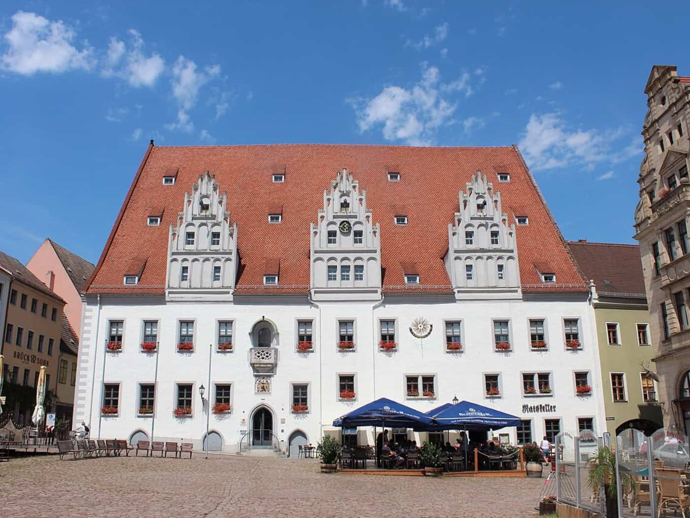 Weißes Rathaus Meißen mit rotem Dach und drei Ziergiebeln am Marktplatz, davor Sonnenschirme eines Lokals