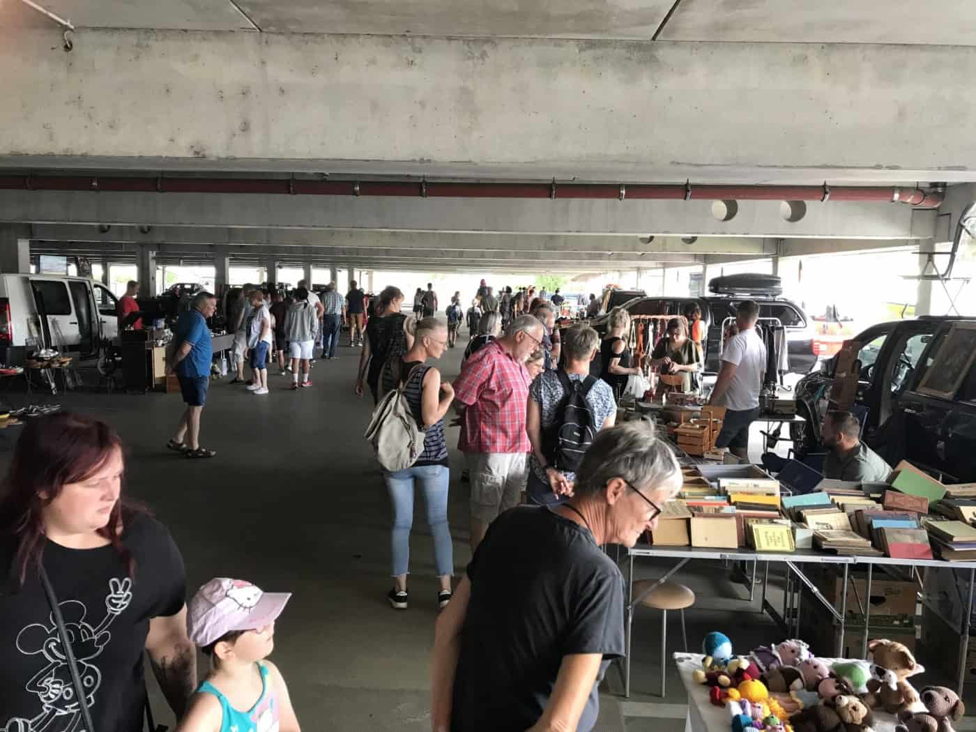 Zahlreiche Menschen stöbern auf einem überdachten Trödelmarkt in einem Parkdeck zwischen Marktständen voller Bücher, Spielzeug und Antiquitäten. Im Vordergrund betrachten Besucher das Angebot an einem Verkaufsstand.