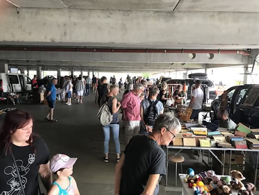Zahlreiche Menschen stöbern auf einem überdachten Trödelmarkt in einem Parkdeck zwischen Marktständen voller Bücher, Spielzeug und Antiquitäten. Im Vordergrund betrachten Besucher das Angebot an einem Verkaufsstand.