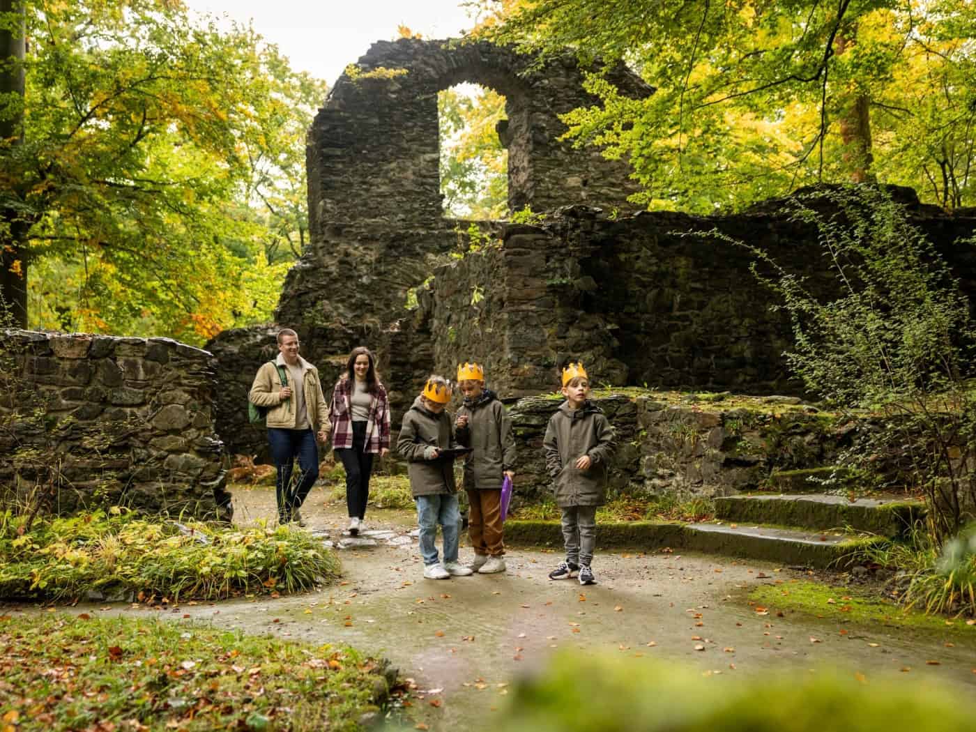 Eine Familie mit drei Kindern wandert an einem Herbsttag durch eine historische Steinruine im Wald. Die Kinder tragen gelbe Papierkronen und erkunden aufmerksam den Weg, während bunte Herbstblätter die verfallenen Mauern umrahmen.   