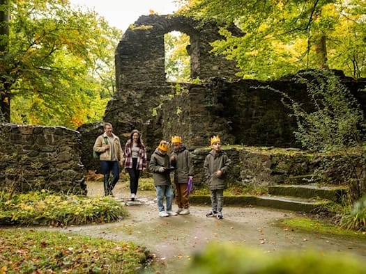 Eine Familie mit drei Kindern wandert an einem Herbsttag durch eine historische Steinruine im Wald. Die Kinder tragen gelbe Papierkronen und erkunden aufmerksam den Weg, während bunte Herbstblätter die verfallenen Mauern umrahmen.   