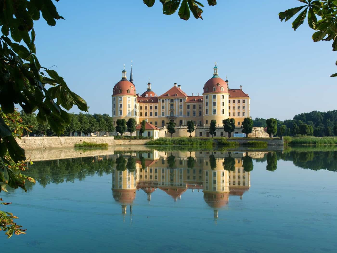 Das Barockschloss Moritzburg spiegelt sich klar im ruhigen Wasser des Schlossteichs unter blauem Himmel. Rahmendes Laub von Kastanienbäumen führt den Blick auf die ockerfarbene Fassade mit den rotdachigen Türmen des Jagdschlosses.