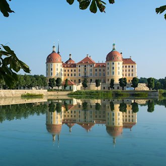 Das Barockschloss Moritzburg spiegelt sich klar im ruhigen Wasser des Schlossteichs unter blauem Himmel. Rahmendes Laub von Kastanienbäumen führt den Blick auf die ockerfarbene Fassade mit den rotdachigen Türmen des Jagdschlosses.
