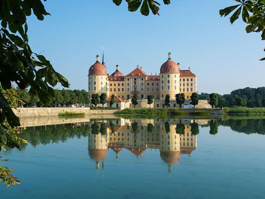 Das Barockschloss Moritzburg spiegelt sich klar im ruhigen Wasser des Schlossteichs unter blauem Himmel. Rahmendes Laub von Kastanienbäumen führt den Blick auf die ockerfarbene Fassade mit den rotdachigen Türmen des Jagdschlosses.