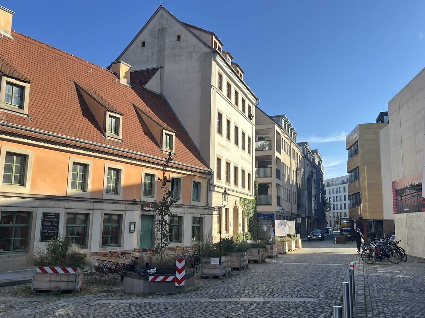 Gepflasterte Altstadtstraße mit historischen Gebäuden; an der Fassade des Umweltzentrums leuchten die Schriftzüge „Arnica montana” und „Sturnus vulgaris”, am Fahrbahnrand stehen Holzhochbeete.