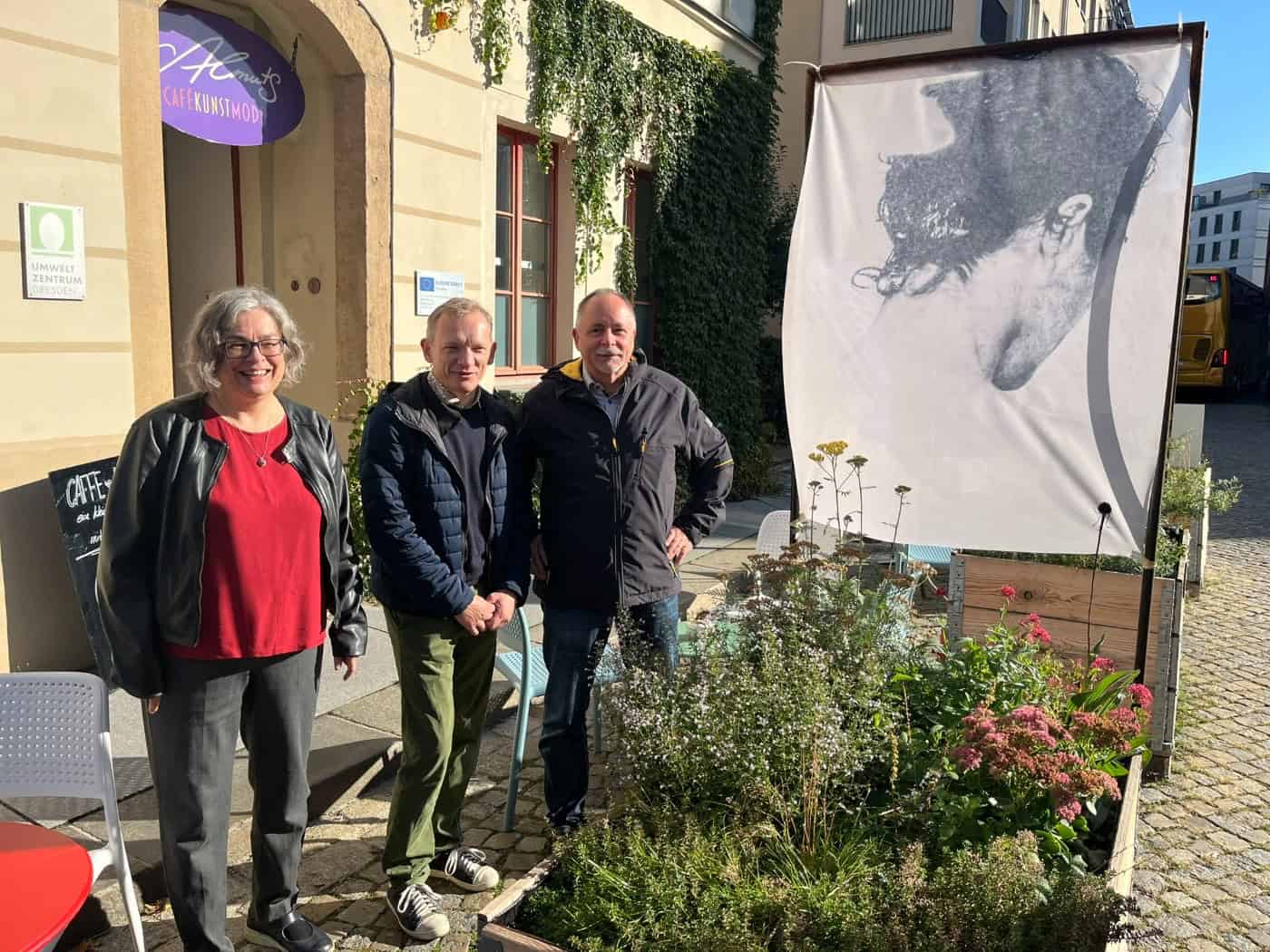 Drei Personen – Eva Jähnigen, Tom Umbreit und André Barth – stehen lächelnd vor einem bepflanzten Holzhochbeet und einem Kunstbanner neben dem Eingang des Umweltzentrums Dresden.